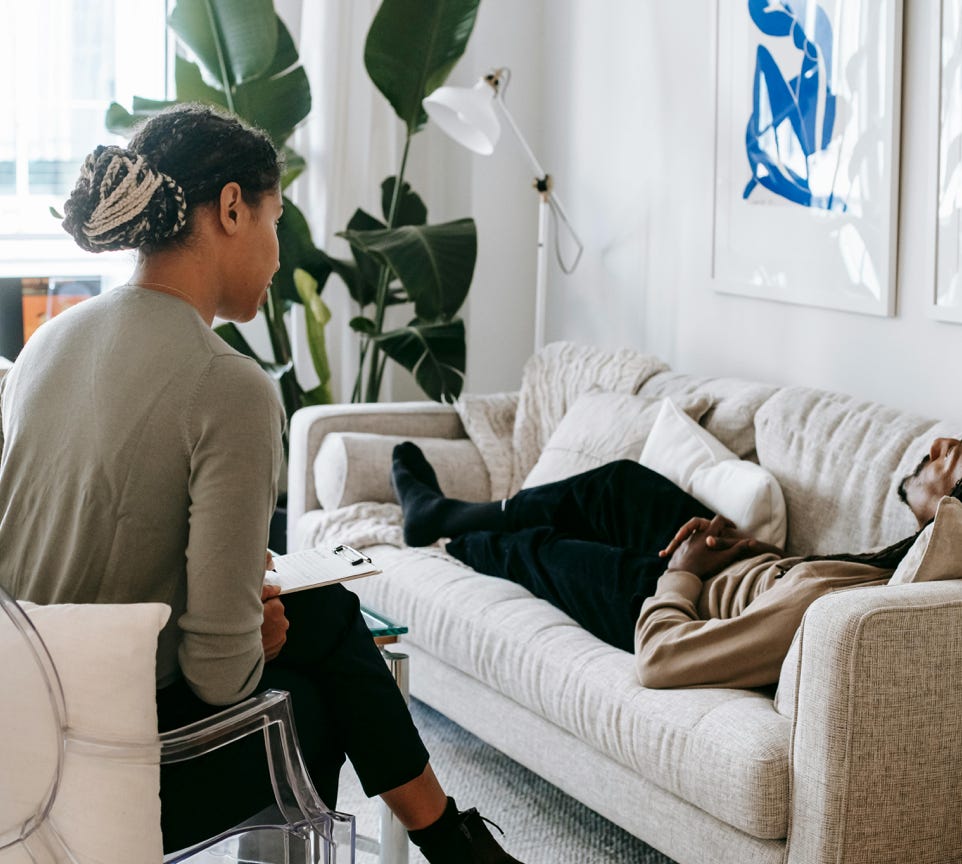 Ethnic female therapist with clipboard sitting near male client lying on couch with clasped hands during psychotherapy session in cozy office